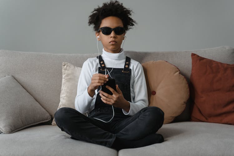 A Child Wearing Sunglasses Holding A Smartphone With Earphones