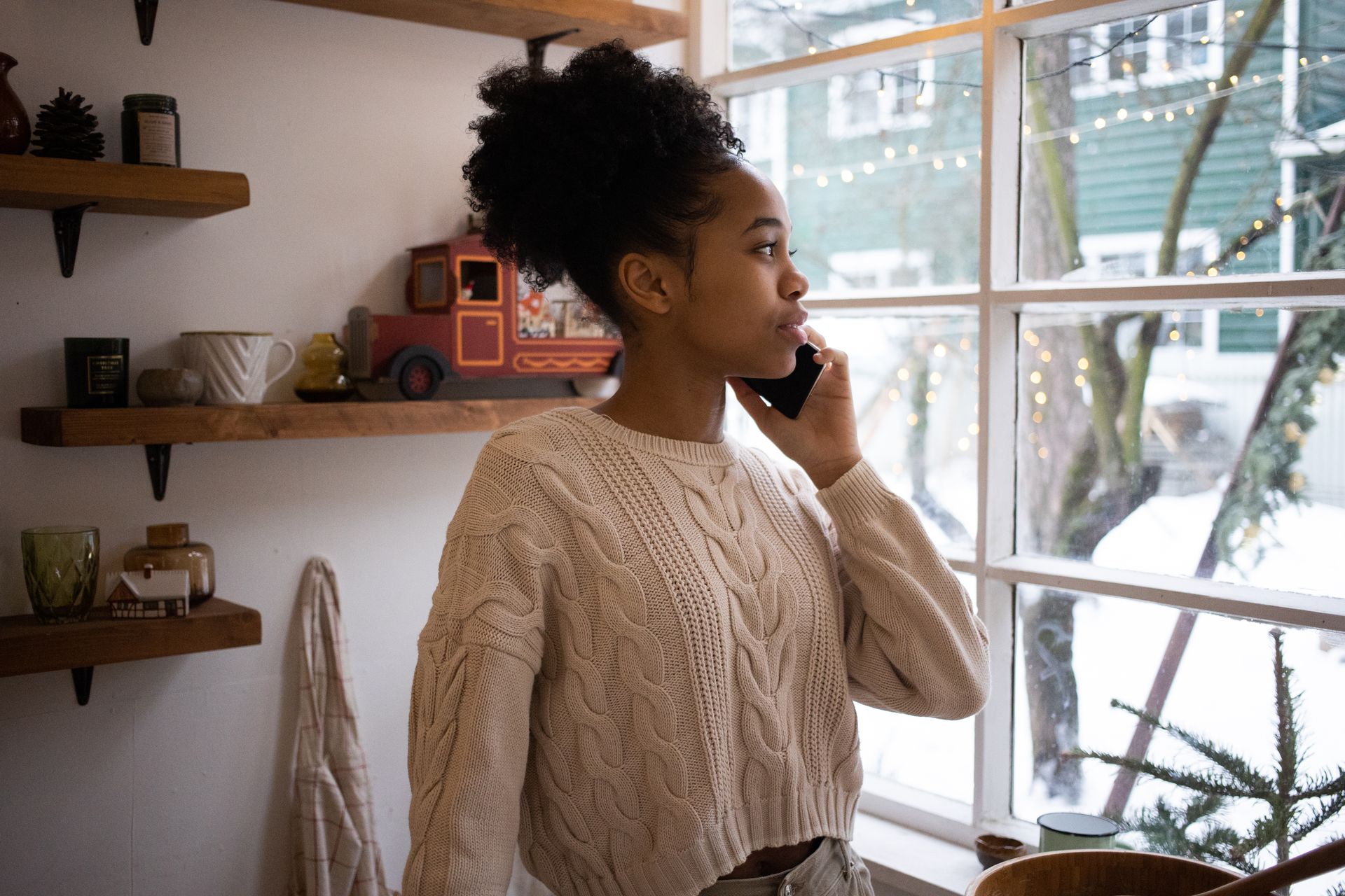 African American woman talking on smartphone by window, cozy winter indoor scene.