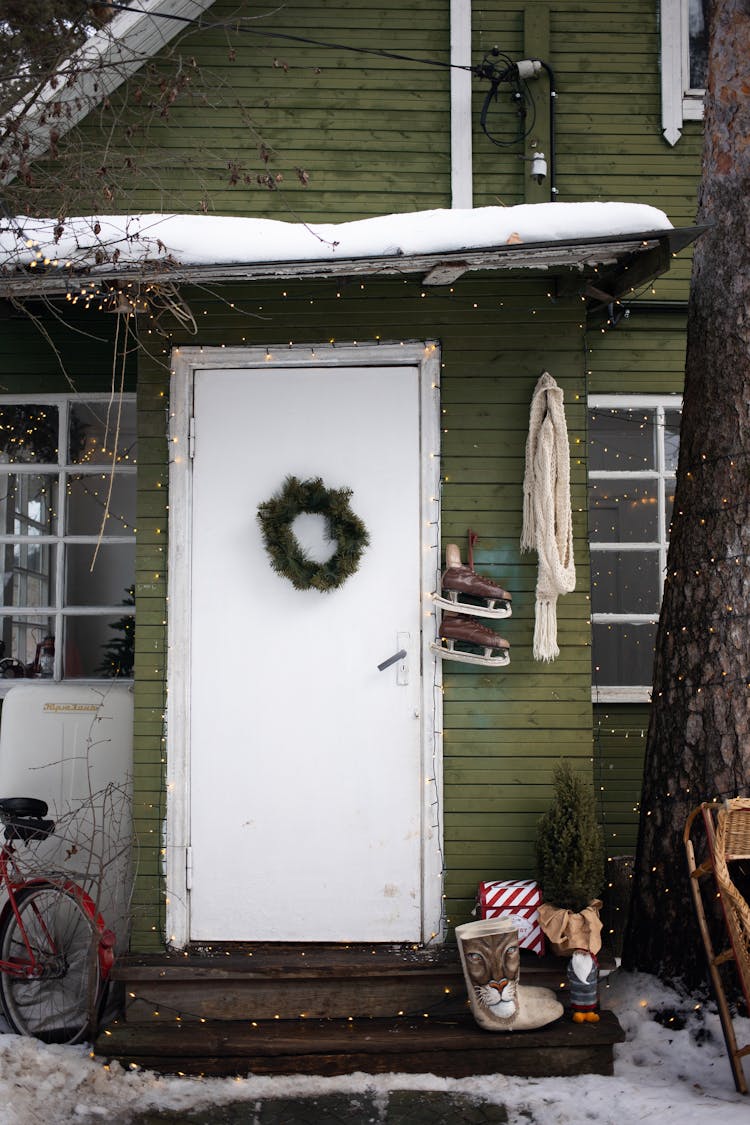 A Christmas Wreath Hanging On A Door Of A Wooden House Decorated With String Of Lights