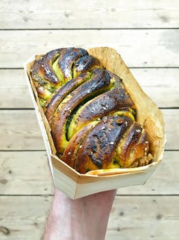 High angle of crop unrecognizable male with bread loaf in hand decorated with pesto and seeds in container near wooden floor in light place
