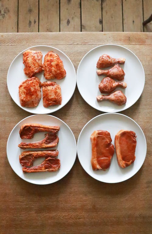 Top view of uncooked different meat fillet and steaks with drumsticks and ribs with seasoning on white plates placed on wooden table