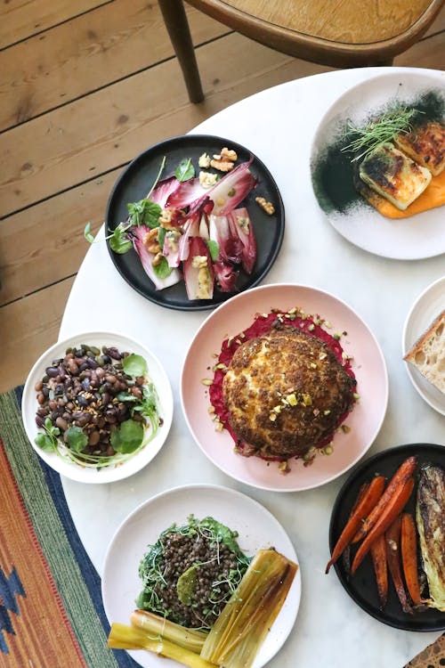 Top view of plates with whole roasted cauliflower near pickled stems with herbs and buckwheat porridge near beans and cutted radichio salad near grilled carrot with corn cob on table