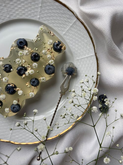 Top view of organic soap decorated with blueberries on white textile with fork placed on white textile near thin branches