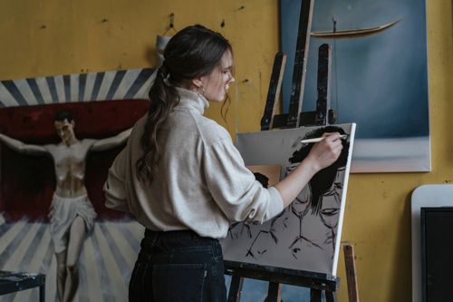 Free Female artist wearing turtleneck painting on canvas with paintbrush. Studio setting with artworks and easel. Stock Photo