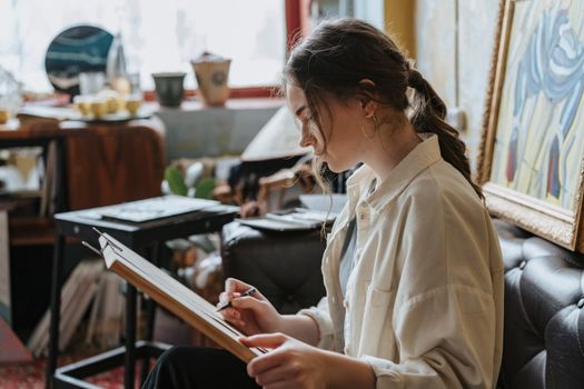 A young woman focuses on her sketching in a cozy, art-filled room, creating a serene mood.