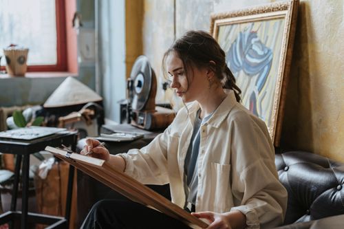 Free A young artist sits on a couch sketching a portrait in a cozy studio setting. Stock Photo