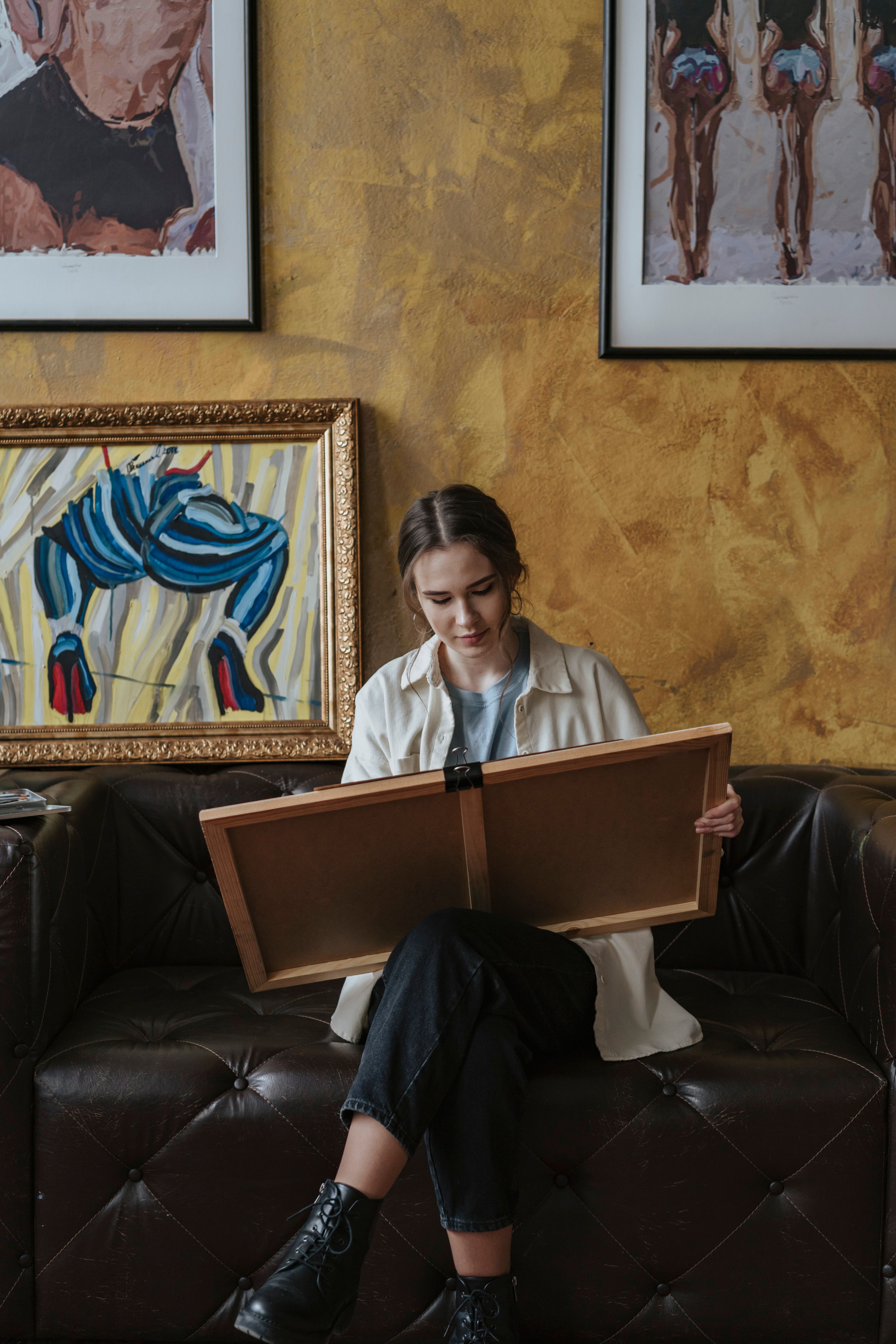 Woman holding a painting while sitting on a sofa under framed artwork.