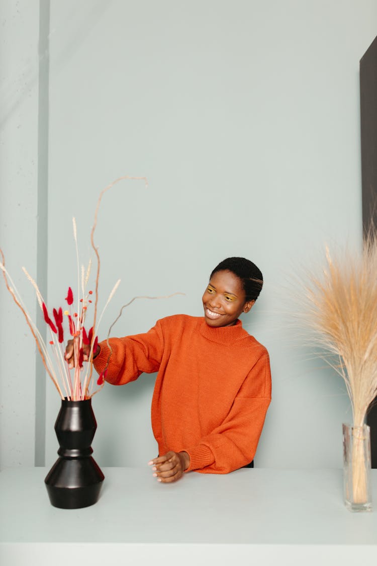 A Woman In Orange Sweatshirt Arranging Flowers On A Black Vase