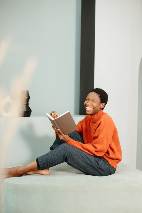 Free Smiling woman in an orange sweater reading a book indoors, creating a cozy and relaxed atmosphere. Stock Photo