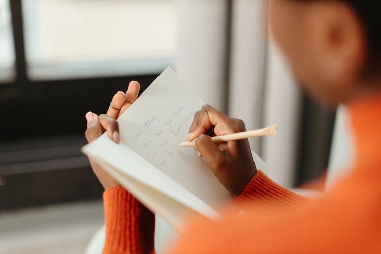 Person In Orange Sweater Writing A Message On A Card Using Pen