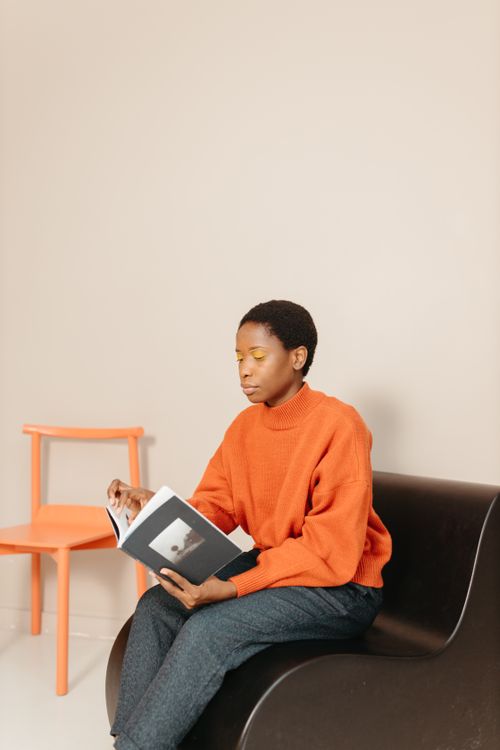 Free A woman in an orange sweater sits on a sofa reading a book with a calm expression. Stock Photo