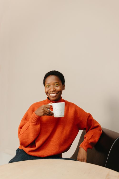 Free Smiling woman in orange sweater holding a mug, seated indoors. Warm and cozy ambiance. Stock Photo