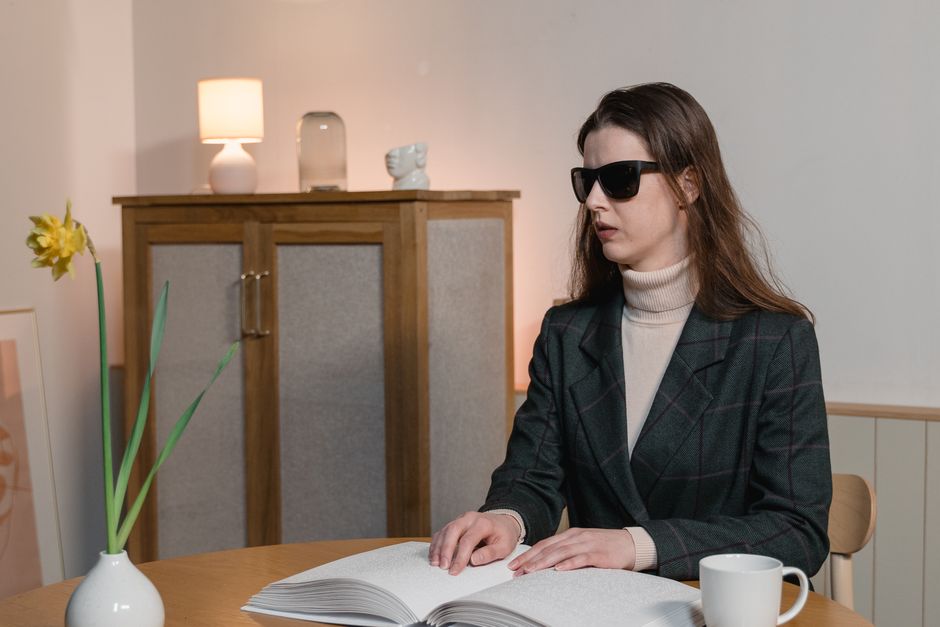 A woman wearing sunglasses reads a braille book at a table, highlighting accessibility and independence.