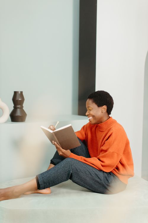 Free Woman sitting indoors, smiling, and writing in a notebook, wearing an orange sweater. Stock Photo
