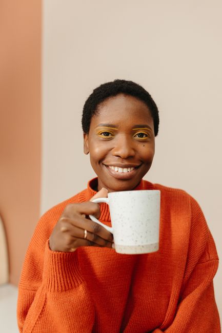 A cheerful woman in an orange sweater holding a mug, expressing warmth and friendliness indoors.