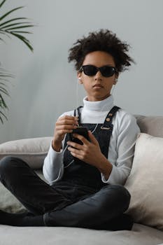 African American girl with afro hairstyle sitting on sofa, listening to music with sunglasses and smartphone.