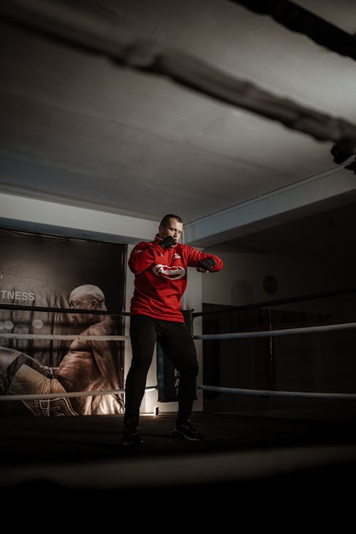 Low angle full body of enduring strong athlete in sportswear standing in middle of boxing ring and training kicks