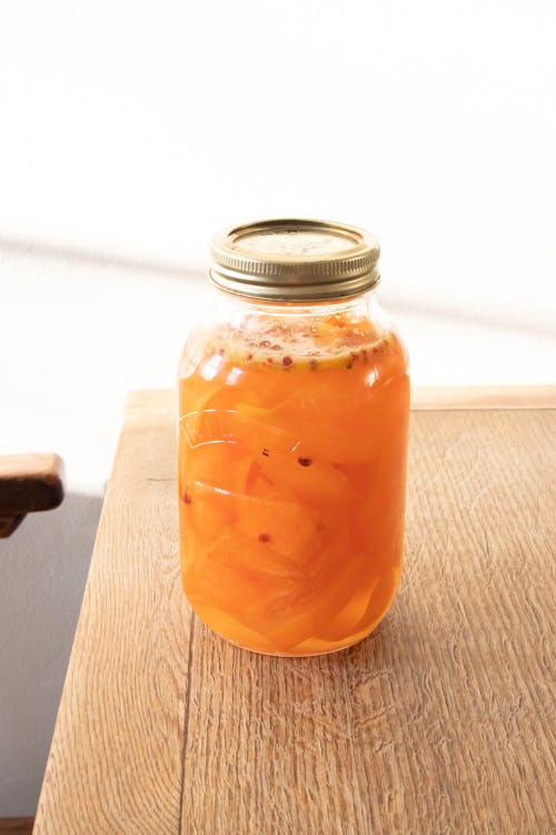 Glass jar of sweet orange canned jam with pieces of fresh fruits placed on wooden table near wall in kitchen