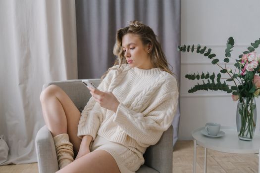 Woman in cozy sweater using smartphone in a modern indoor setting, relaxing on an armchair.