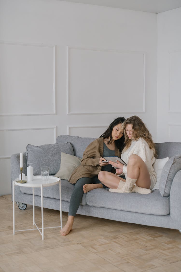 Women Wearing Sweaters Sitting Together On A Sofa Reading A Book