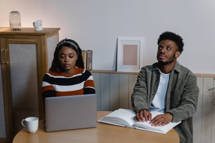 A Woman Using A Laptop Sitting Beside A Man Reading A Braille Book