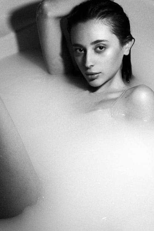 Black and white of sensual young female with dark wet hair lying in bathtub with foam and looking at camera