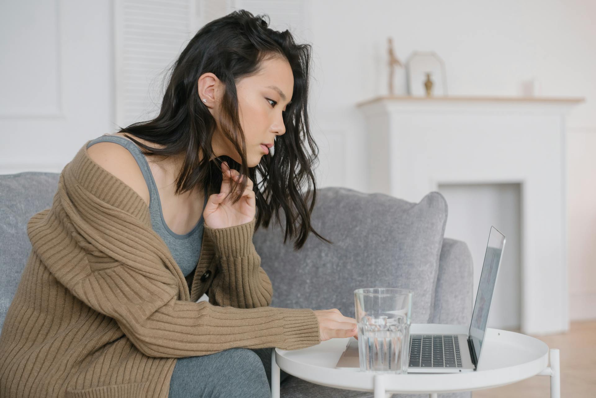 A Woman in Brown Cardigan Typing on Her Laptop Beside the Glass of Water