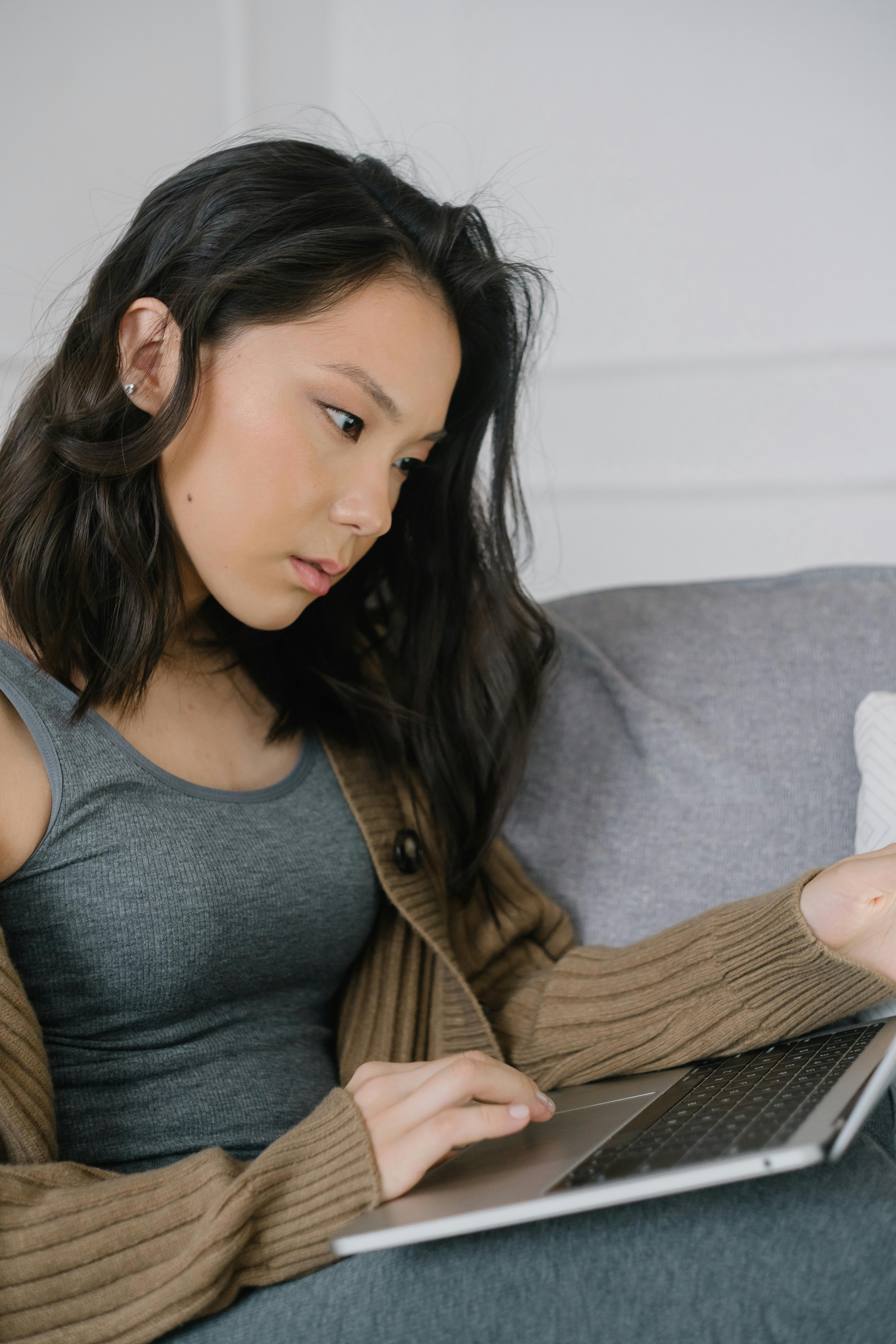 Asian woman intently working on a laptop while relaxing on a cozy sofa.