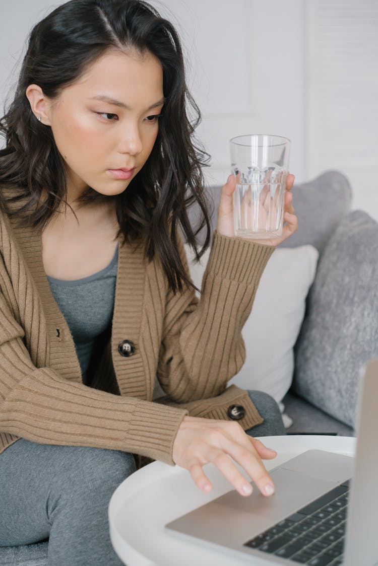 Woman In Brown Cardigan Sitting On Couch Holding A Glass Of Water While Scrolling On Her Laptop