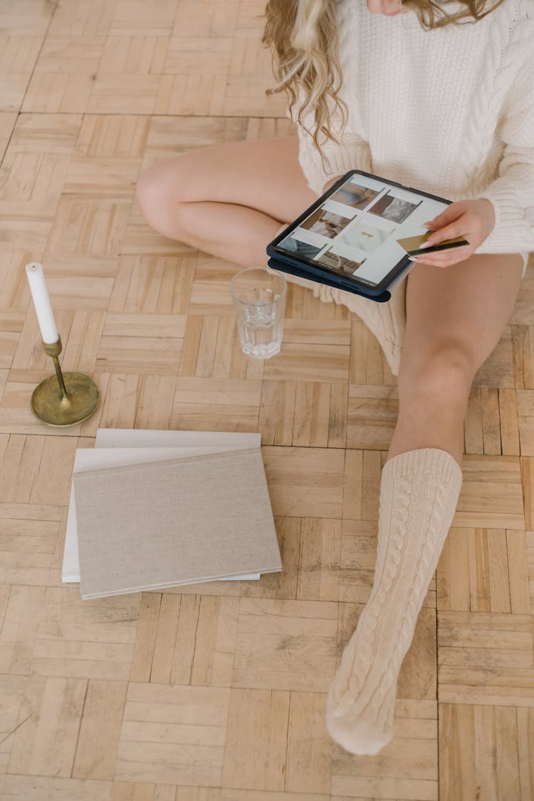 Person In White Sweater Sitting On Wooden Floor Holding An IPad And Credit Card