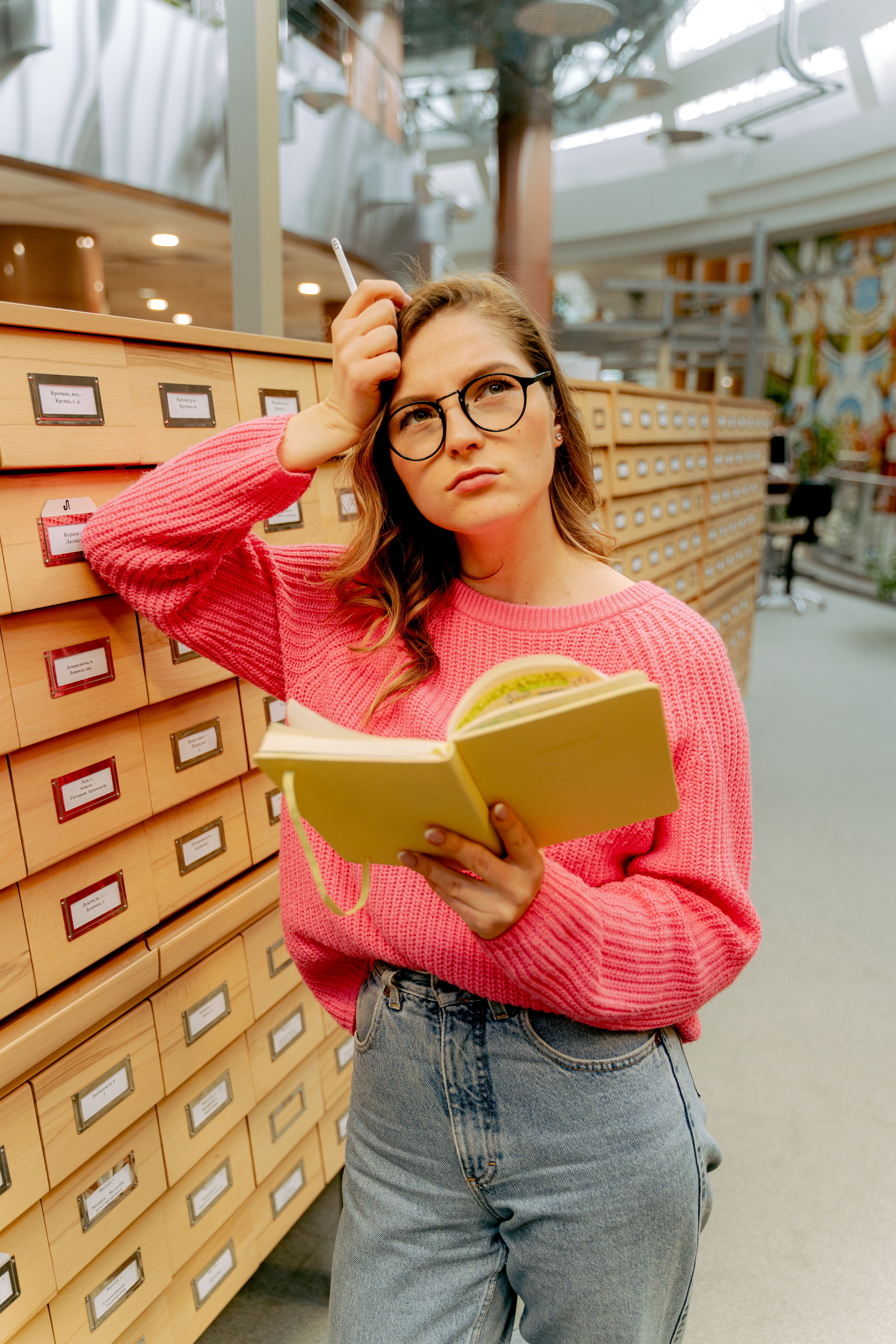 Young woman in glasses reading a book in a library. Contemplative expression next to card catalogs.