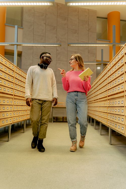Free Two diverse students walking and talking in a library aisle with card catalogs. Stock Photo