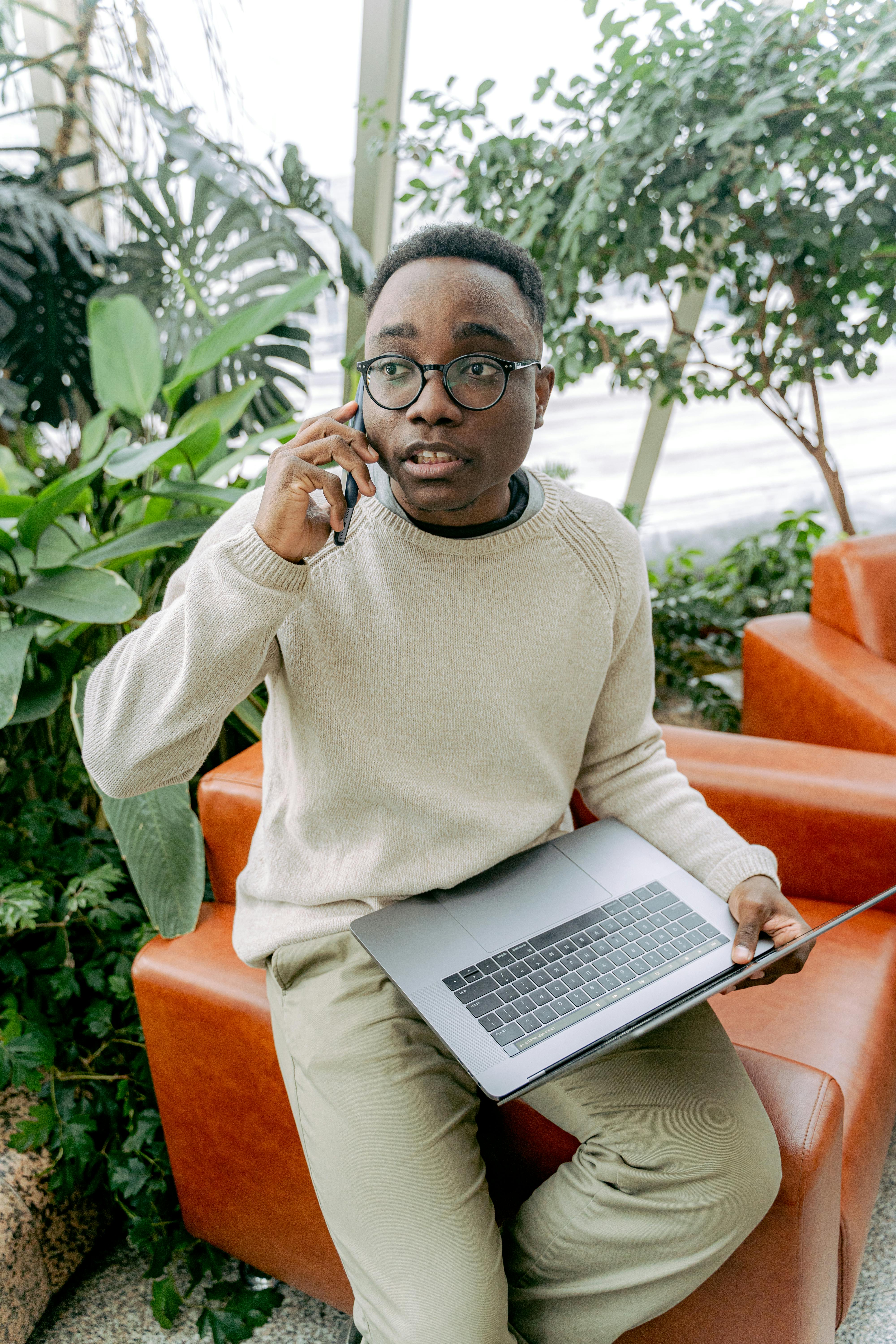 A young man in an indoor setting, sitting with a laptop and talking on a mobile phone.