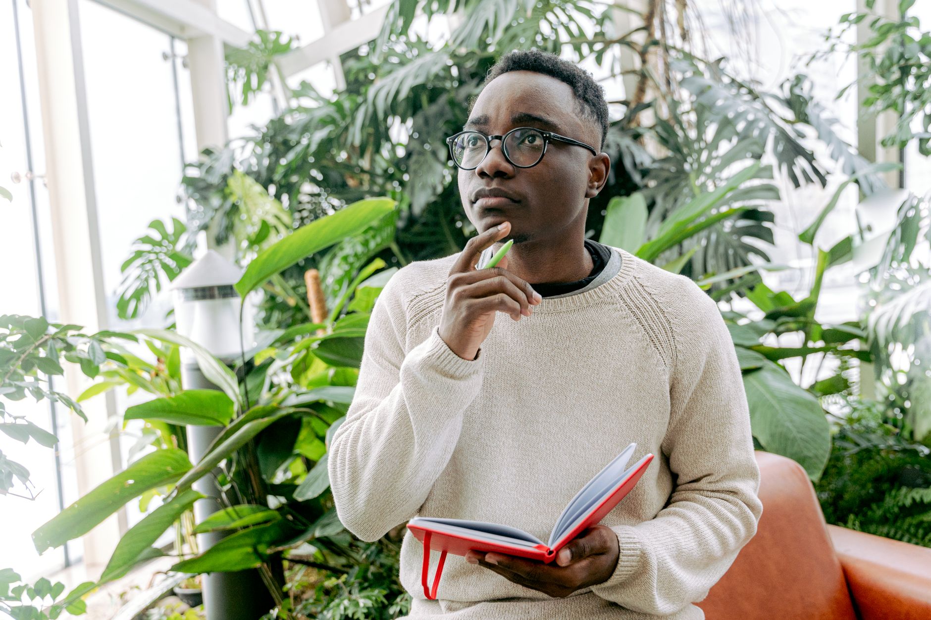 A man with eyeglasses holding a notebook, pondering in a lush indoor garden.