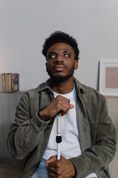 Man with visual impairment holding a cane, seated indoors, contemplative expression.