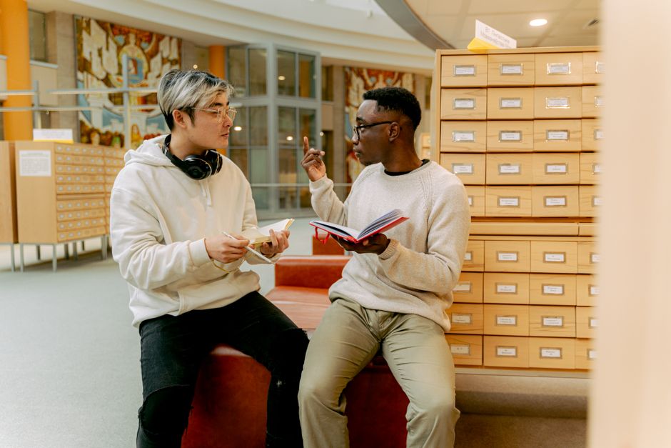 Two young men discussing in a library, exchanging ideas and holding notebooks. Two young men discussing in a library, exchanging ideas and holding notebooks.