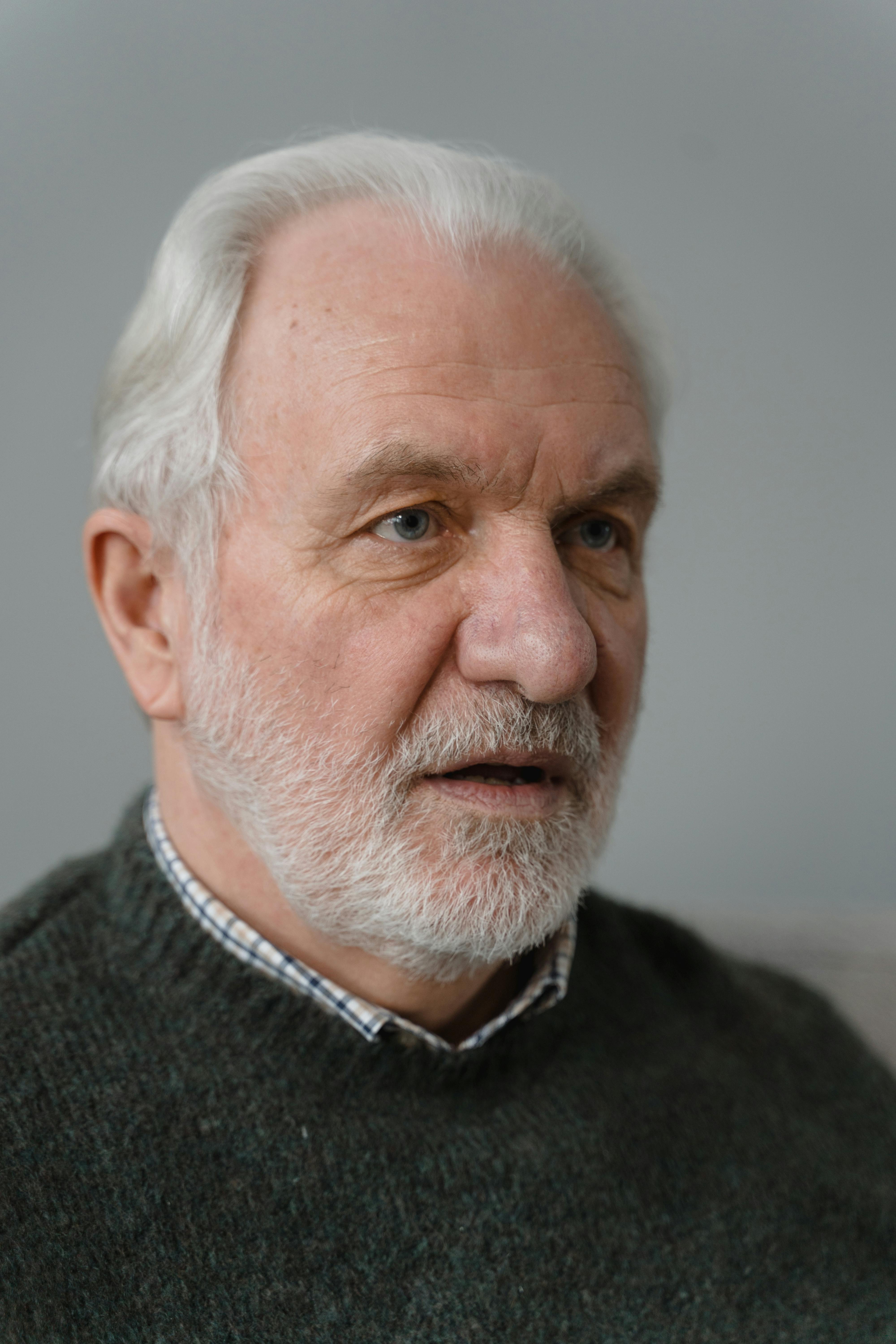Close-up portrait of an elderly man with a gray beard and thoughtful expression against a neutral background.