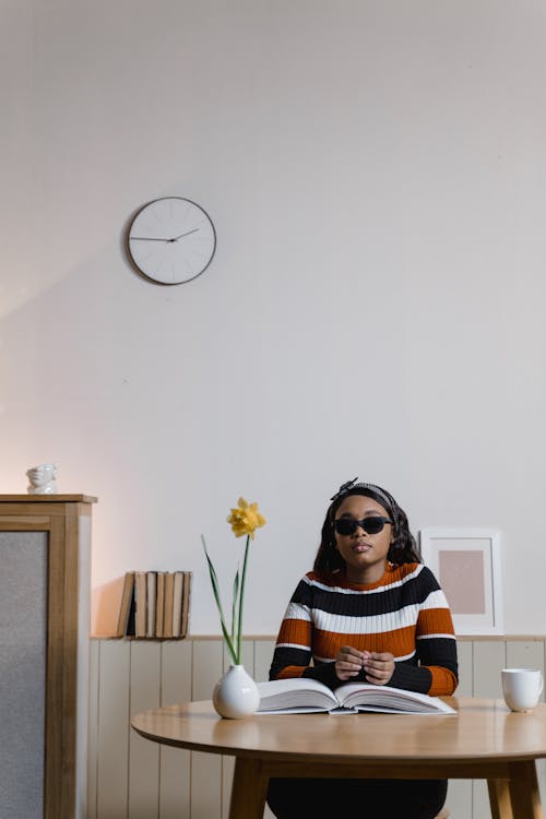 A Woman in Striped Knitted Sweater Wearing Sunglasses while Sitting Near the Wooden Table with Braille Book