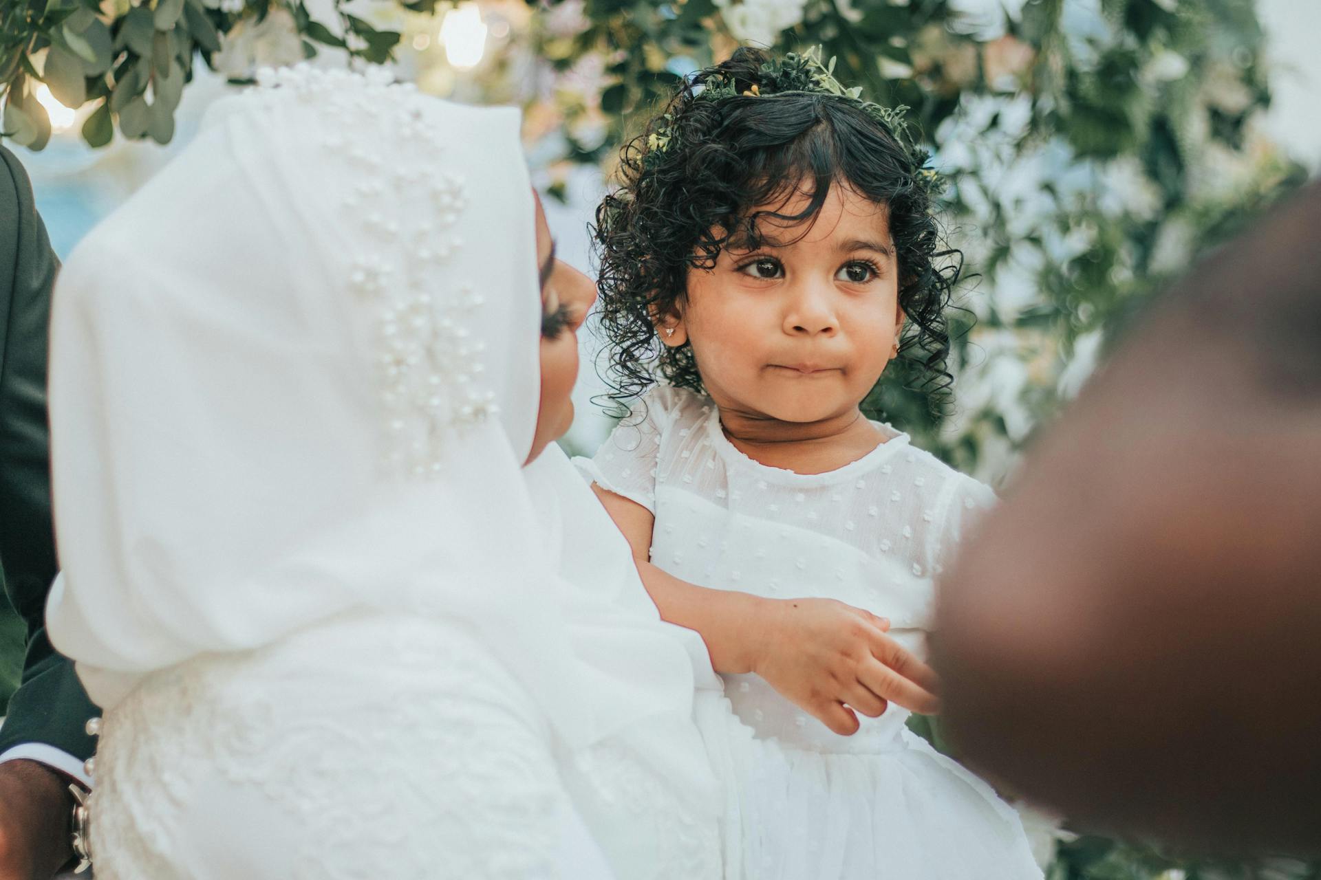 Indian bride in traditional wedding outfit looking at Indian daughter in hands while standing near green plants during holiday celebration