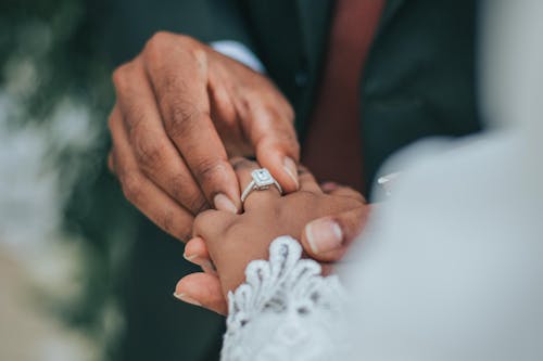 Crop faceless groom putting wedding ring on finger of anonymous bride in white wear against blurred background during wedding ceremony