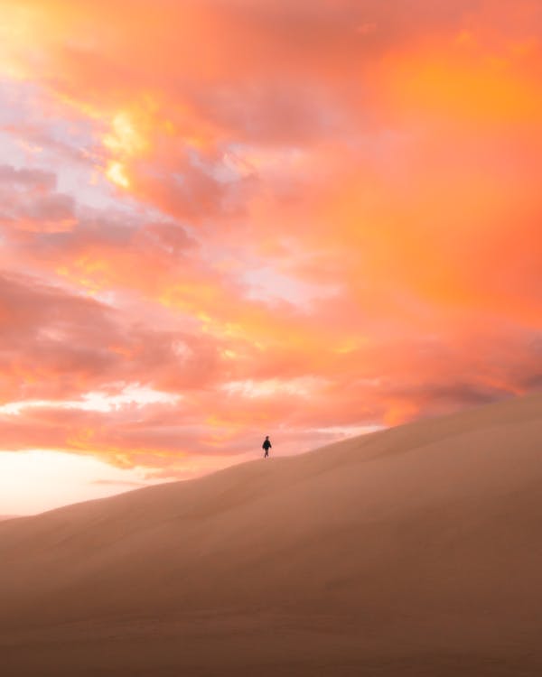 Anonymous traveler walking on sandy dune in endless desert against picturesque cloudy sunset sky
