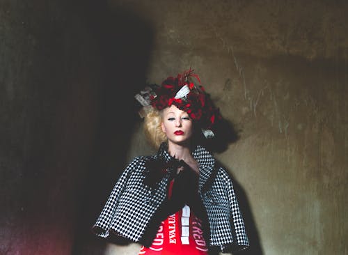 Serious young female with bond hair and bright makeup in elegant outfit and creative headdress standing against shabby wall and looking at camera