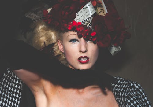 Self assured young female model with blond hair and red lips in creative floral headdress touching chin and looking at camera against black background