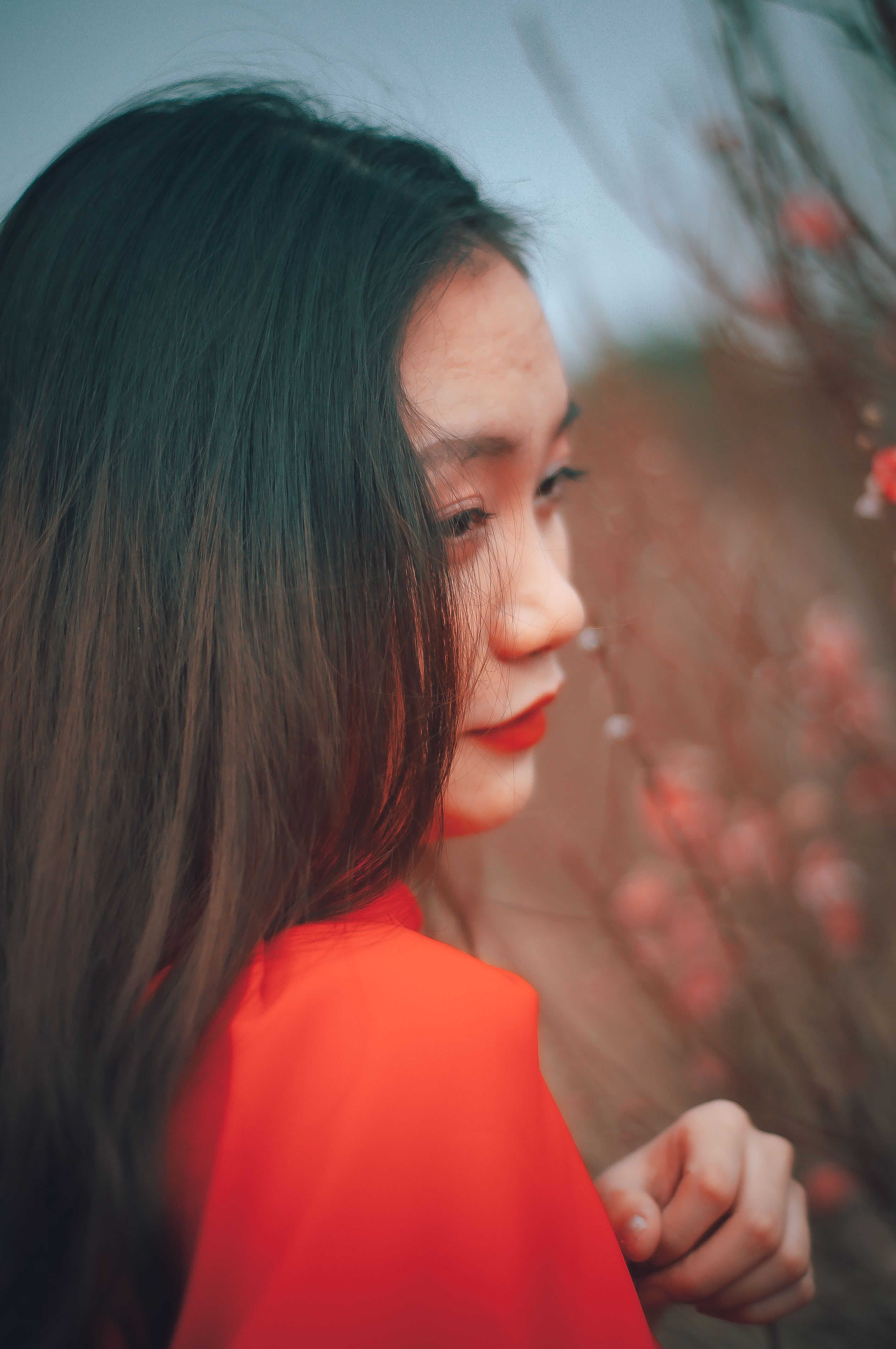 A thoughtful woman in a red dress poses outdoors among blooming flowers.