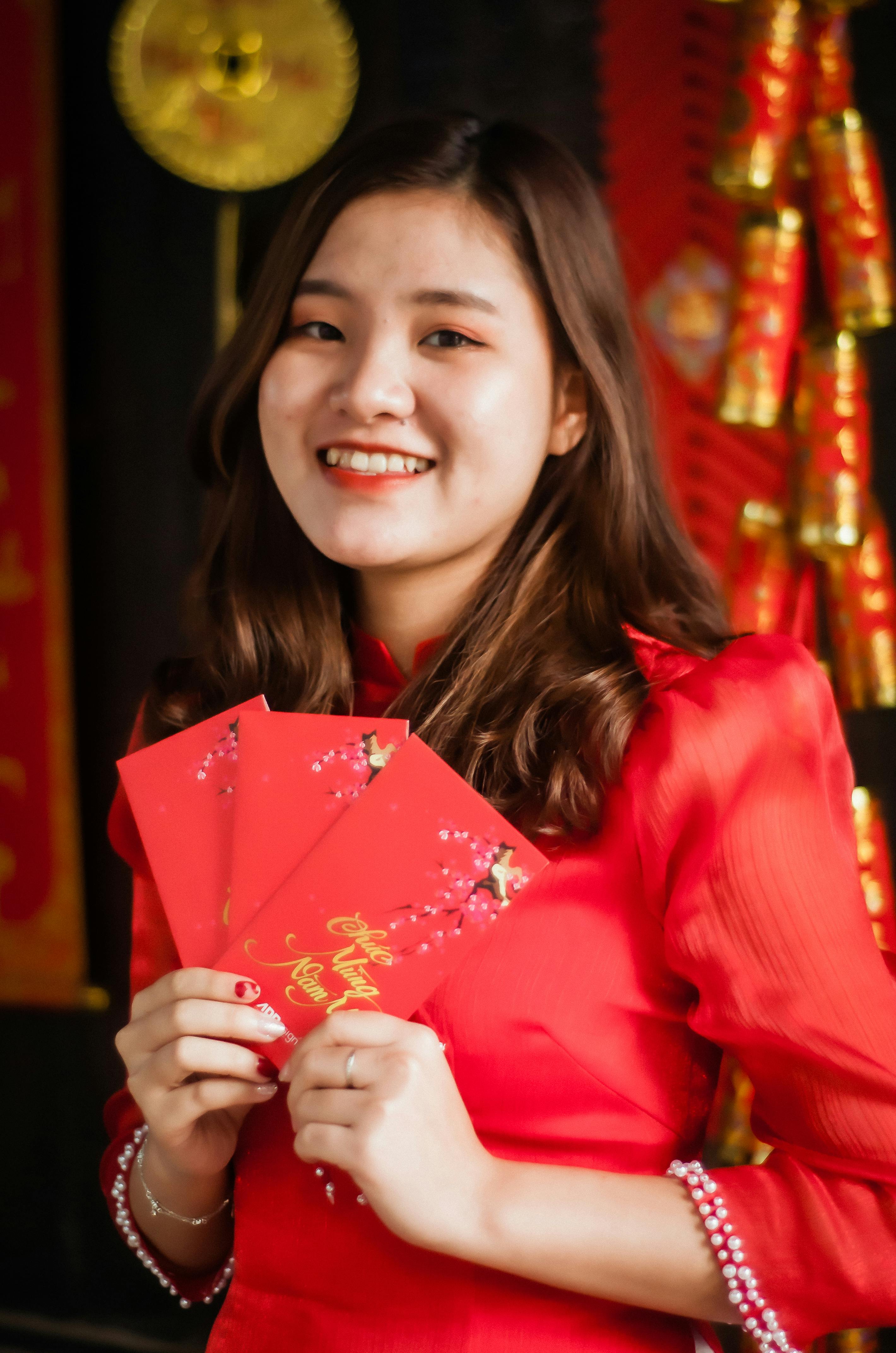 Smiling Asian woman in traditional attire holding festive red envelopes for Lunar New Year celebration.