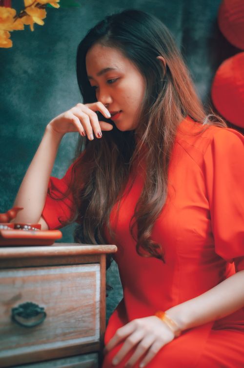 Free Asian woman in red dress sitting thoughtfully by a wooden drawer with decorative elements. Stock Photo