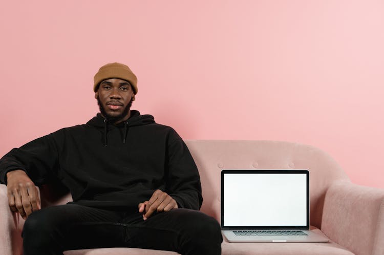 A Man Sitting On A Couch Beside A Laptop With Blank Screen