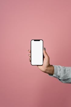 Close-up of a hand holding a smartphone with a blank screen against a pink backdrop.