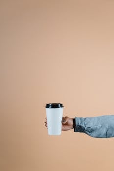 Person's hand holding a disposable coffee cup against a beige background.