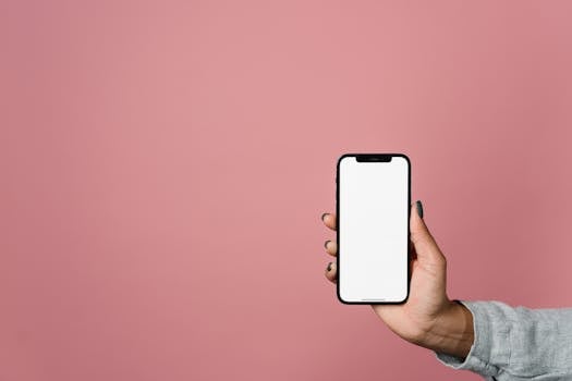 A hand holds a blank-screen smartphone against a solid pink background, perfect for mockups.